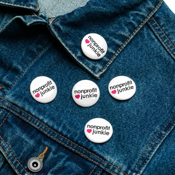 Set of white pin buttons, black text on one side says nonprofit junkie in regular arial like font next to a pink heart, on a denim jacket