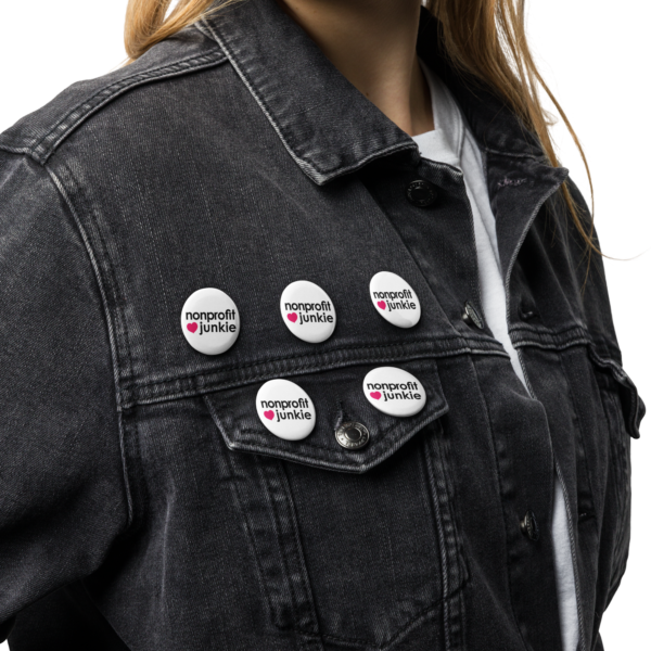 Set of white pin buttons, black text on one side says nonprofit junkie in regular arial like font next to a pink heart, on a denim jacket